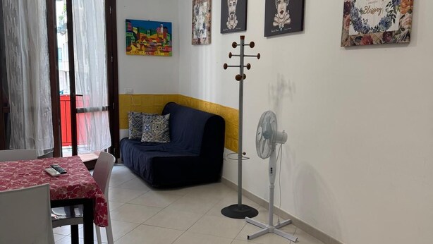 A living room with a sofa, a dining table with chairs, a standing fan, and artwork on the walls.
