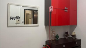 A room with a red and black cabinet, a small table with decorative items, a fire extinguisher, and a mirror with a reflection of a door and a light switch.