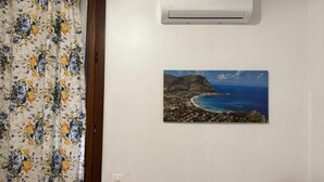 A room with a bed, a desk, a red lamp, a brown ottoman, an air conditioner, and a framed picture of a coastal landscape.