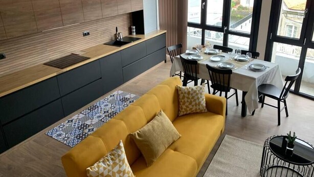 A modern kitchen with a built-in island, dining area with a table set for four, and a yellow sofa with patterned cushions.