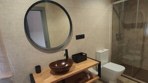 A modern bathroom with a round mirror, a wooden vanity, a brown bowl sink, a toilet, and a glass shower enclosure.