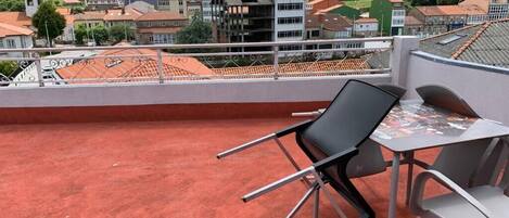 A rooftop terrace with a table and chairs, overlooking a residential area with houses and a hill in the background.