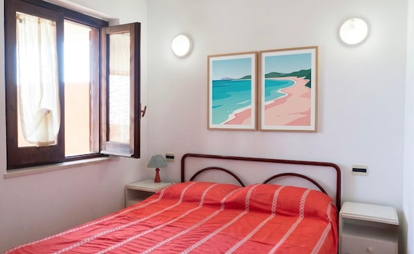 A bedroom with a bed, a nightstand, a lamp, a window with curtains, and two framed pictures on the wall.