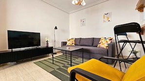 Una sala de estar con televisor de pantalla plana, un sofá con cojines de colores, una mesa de centro, una lámpara de pie y un sillón amarillo.