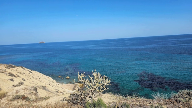 Un paisaje costero con mar azul cristalino, dunas arenosas y vegetación escasa.