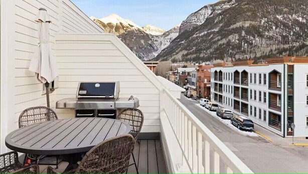 En balkon med grill, fletstole og et bord med udsigt over en gade med parkerede biler og et bjerg i baggrunden.