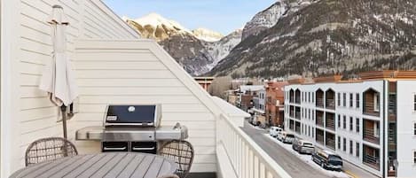 A balcony with a grill, wicker chairs, and a table overlooking a street with parked cars and a mountain backdrop.