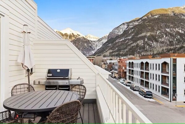 Een balkon met een barbecue, rieten stoelen en een tafel, met uitzicht op een straat met geparkeerde auto's en een berg op de achtergrond.