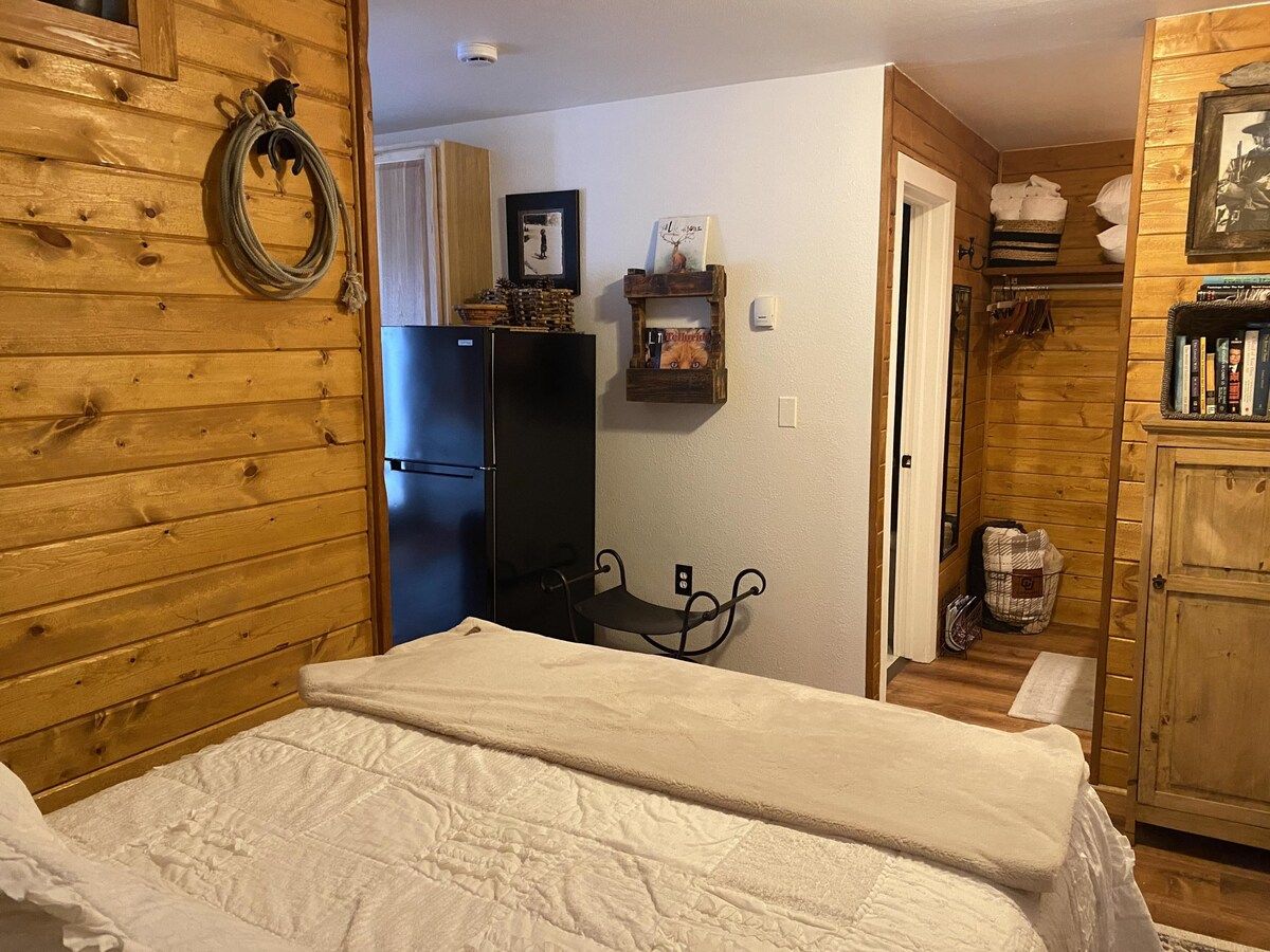 A wooden cabin interior with a bed, a small table, a refrigerator, and a shelf with various items.