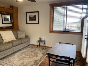 A living room with a sofa, a small table, a patterned rug, and a window with blinds.