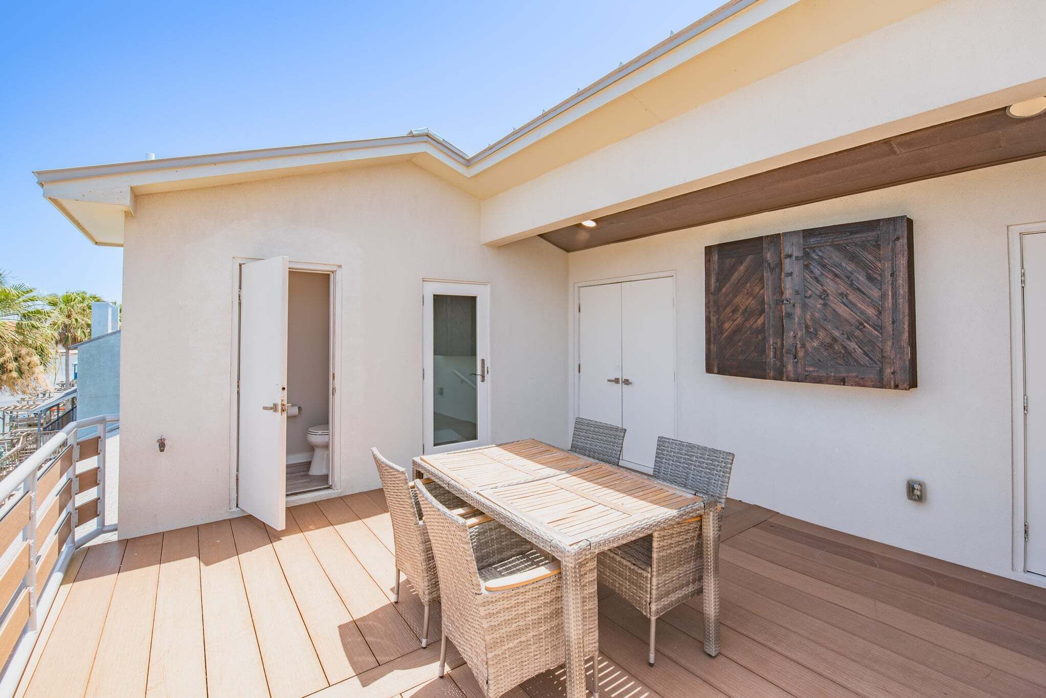 Un patio techado con una mesa de madera y sillas de mimbre, una puerta blanca y una obra de arte con marco de madera en la pared.
