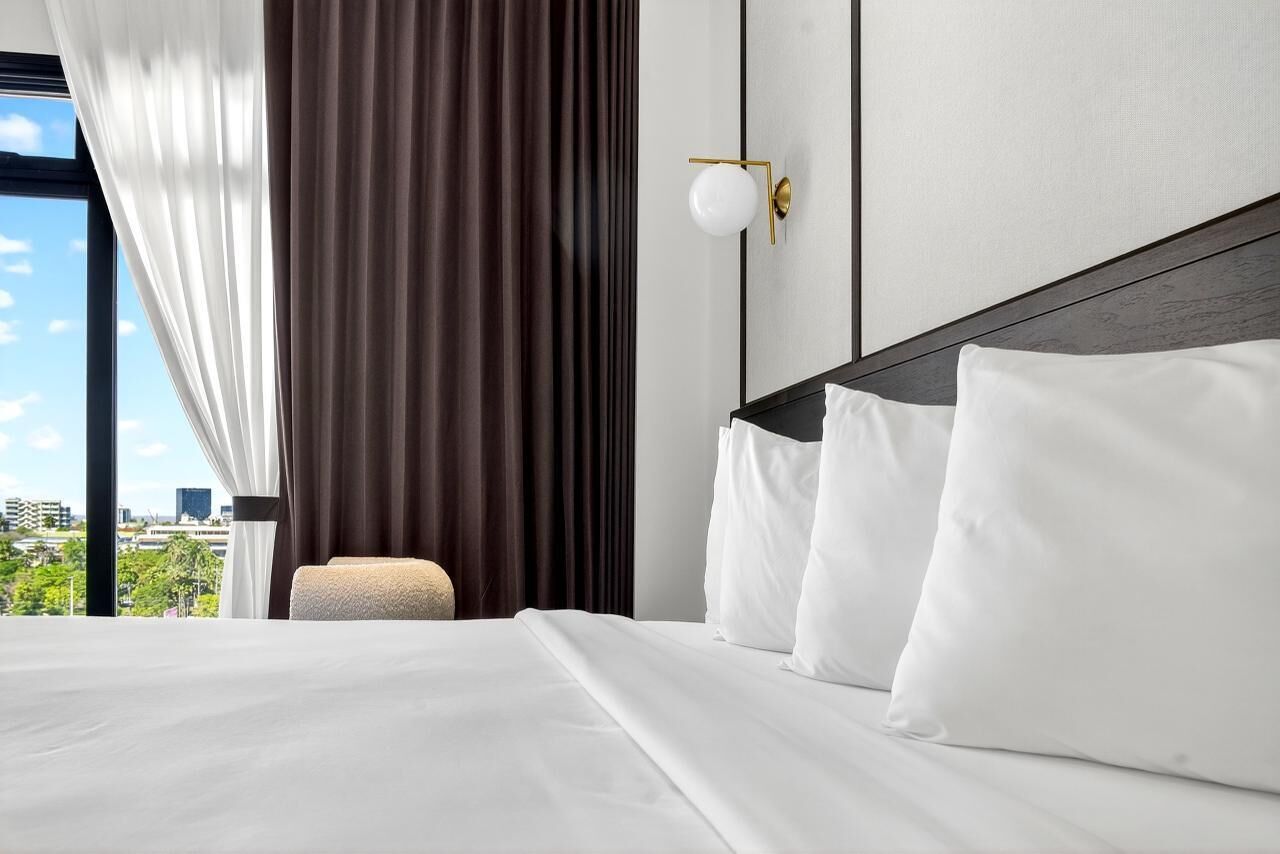 A hotel room with a large bed, white linens, a view of the city, and a modern light fixture.