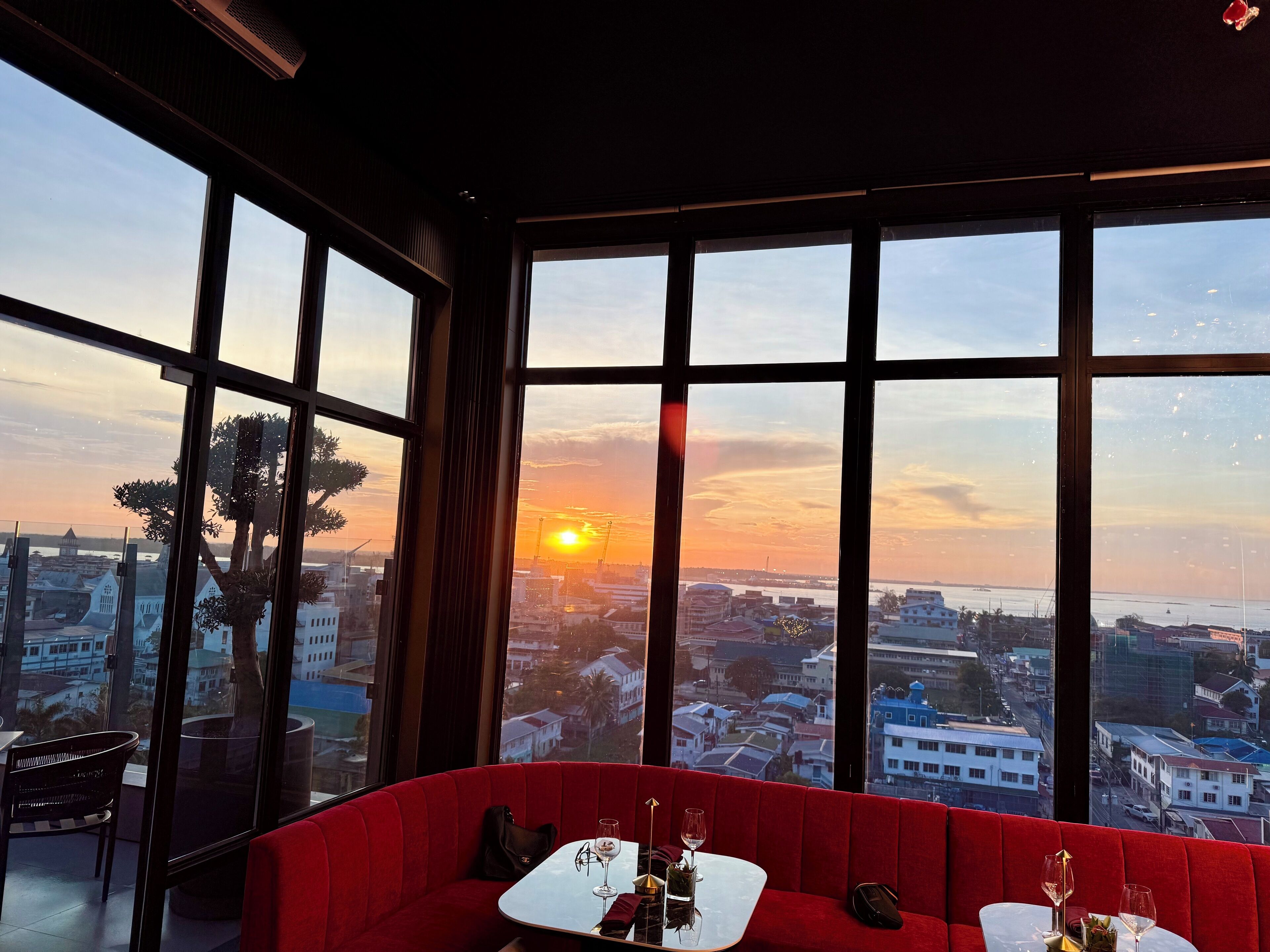 A red upholstered seating area with a table set for two, overlooking a cityscape at sunset.