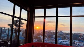 A red upholstered seating area with a table set for two, overlooking a cityscape at sunset.