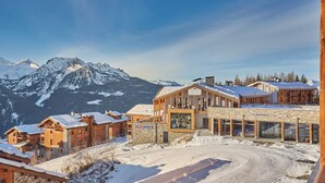 Un chalet de montagne à l’architecture en bois, avec un télésiège et des montagnes enneigées en arrière-plan.