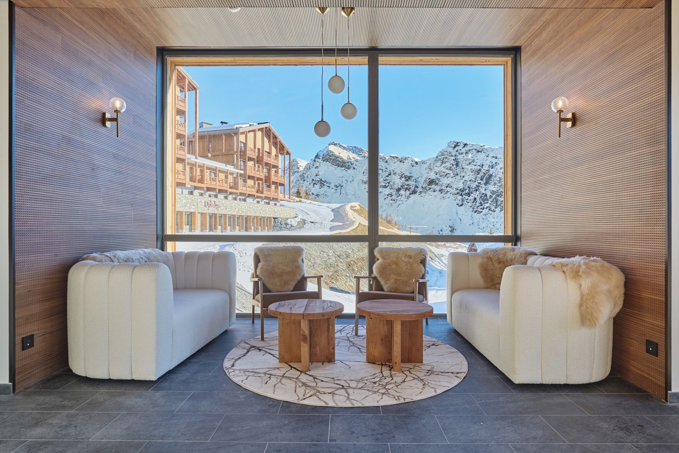 Ein modernes Interieur mit einem großen Fenster, das einen Blick auf einen verschneiten Berg und ein Gebäude freigibt. In der Mitte befinden sich zwei Sofas und zwei Holztische.