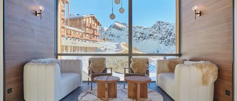 Un interior moderno con un gran ventanal que ofrece vistas a una montaña nevada y un edificio. Hay dos sofás y dos mesas de madera en el centro.
