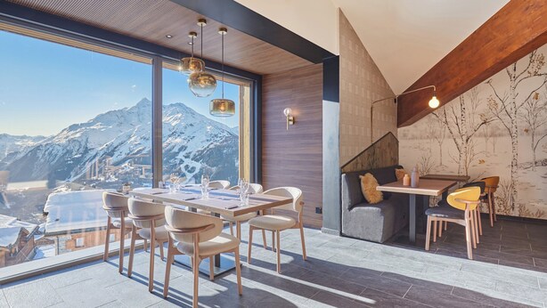 Un espace repas moderne doté d’une grande fenêtre donnant sur des montagnes enneigées, de meubles en bois et d’un coin salon confortable avec une cheminée.