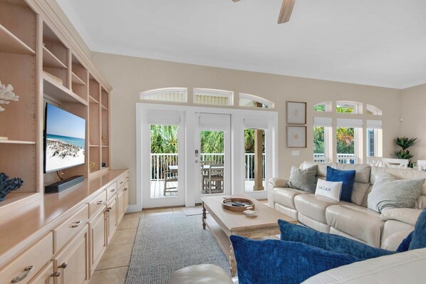 A living room with a beige sofa, a coffee table, and a TV on a wooden cabinet. There is a sliding glass door leading to an outdoor patio with a view of greenery.
