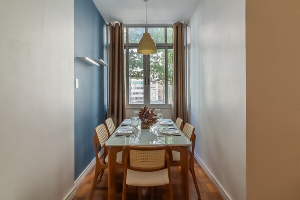 A dining area with a glass table set for six, surrounded by wicker chairs, a hanging light fixture, and a window with curtains. - 2 Minutes From Copacabana Beach D302 (Rio de Janeiro)