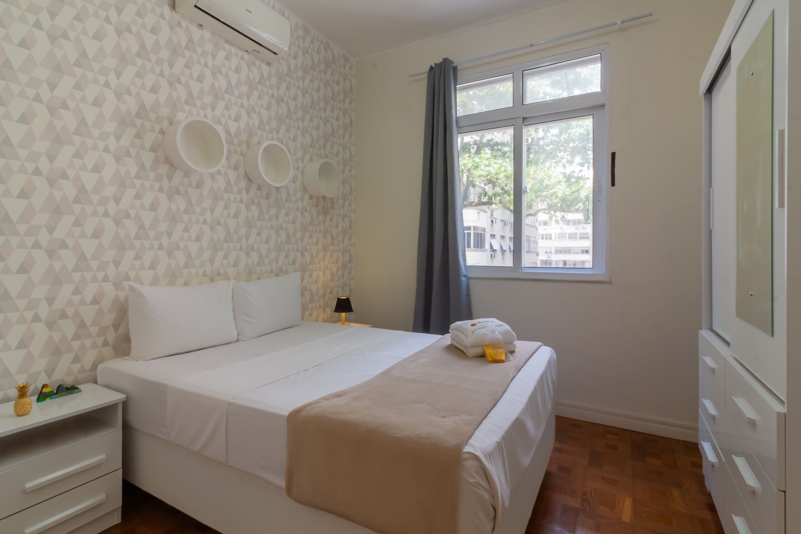 A neatly arranged hotel room with a bed, a nightstand, a dresser, and a window with a view of buildings outside.