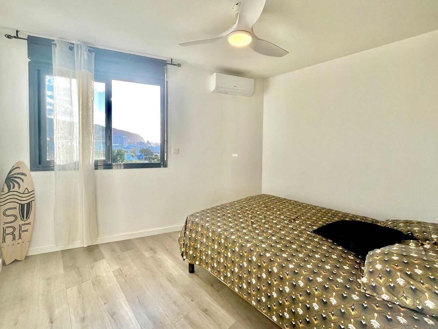 Een slaapkamer met een bed, een plafondventilator, een airconditioning en een raam met uitzicht op gebouwen.