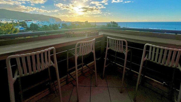 A terrace with a view of the ocean and a sunset. There are three white plastic chairs and a wooden table.