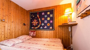 Ein Zimmer mit Holzwandverkleidung, ausgestattet mit einem Bett, einer Steppdecke, Wandteppich, einer Lampe und einem Bücherregal.