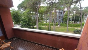 Un balcon donnant sur un espace paysager, comprenant des arbres et un immeuble doté d’une échelle de secours. On y trouve deux chaises en bois.