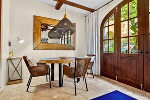 Un comedor con una mesa redonda, cuatro sillas de mimbre, una lámpara colgante, una puerta de madera con paneles de vidrio y un espejo en la pared.