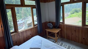 Una habitación con una cama, una mesita con un ventilador y ventanas amplias que dan a un paisaje verde.