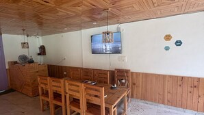 Una habitación con techo de madera, un televisor montado en la pared, una mesa de madera con sillas y suelo de piedra.