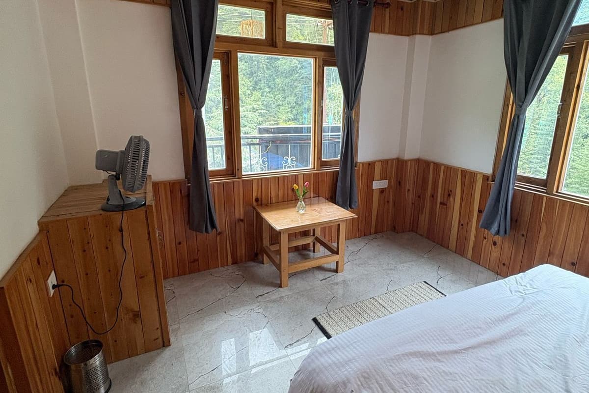 Une pièce aux murs en bois, avec un lit, une petite table sur laquelle est posé un vase, un ventilateur et une fenêtre avec des rideaux.