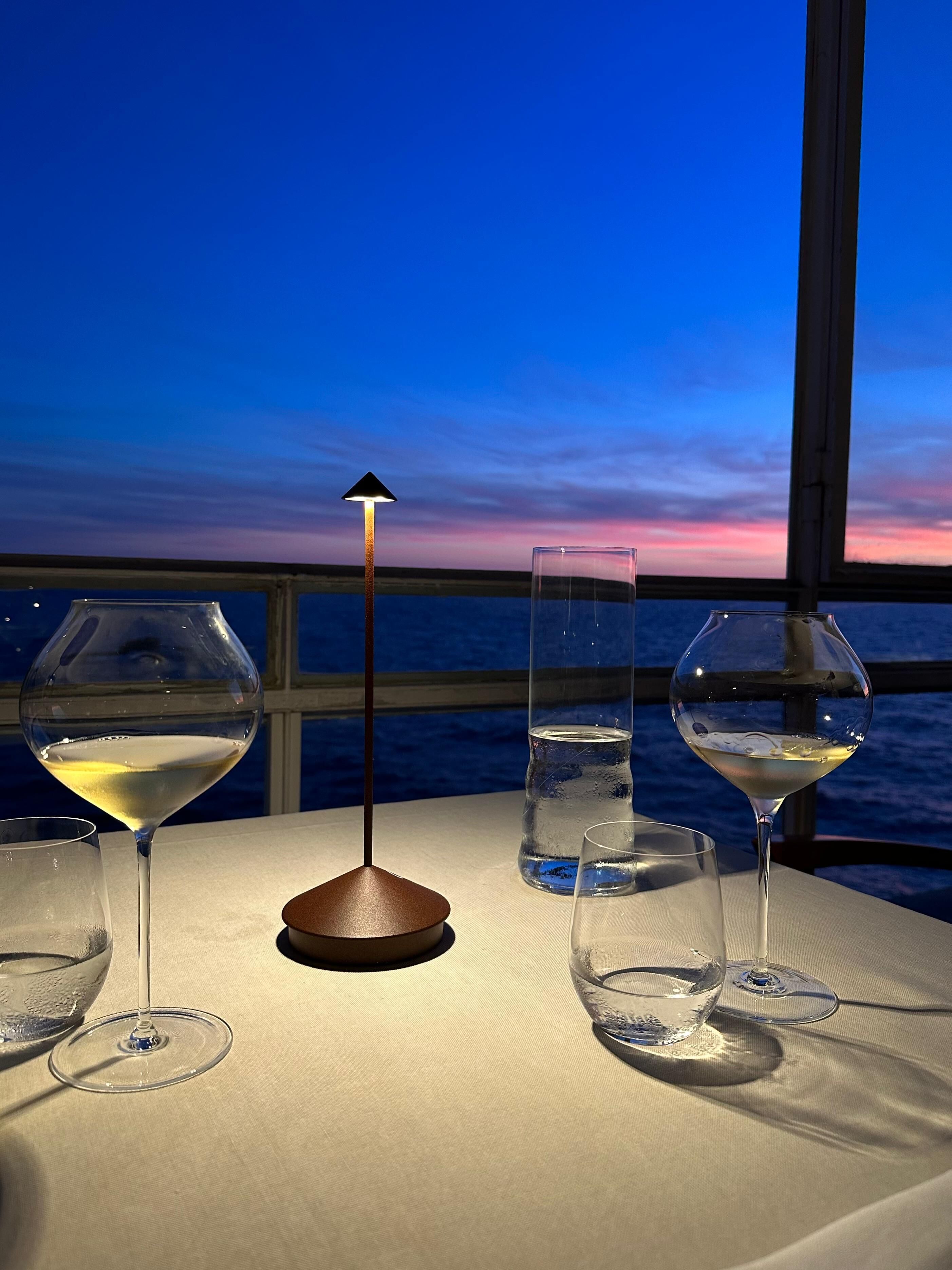 A table with four wine glasses, a tall glass of water, and a floor lamp against a backdrop of a twilight sky.