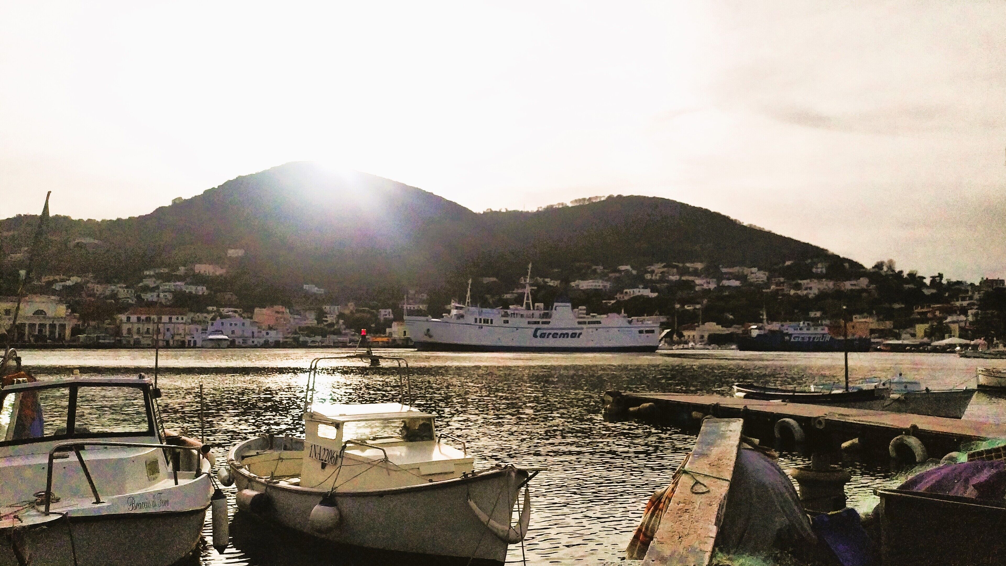 A harbor with boats docked, a large ship named 'Caremar' in the background, and a mountainous landscape.