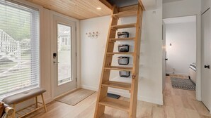 Una estantería de madera con escalones y cestas de almacenamiento frente a una pared blanca, un banco y una puerta que da al exterior.