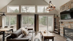 Un salón amplio con sofá de cuero, mesa de centro de madera y chimenea. Grandes ventanales ofrecen vistas al exterior.
