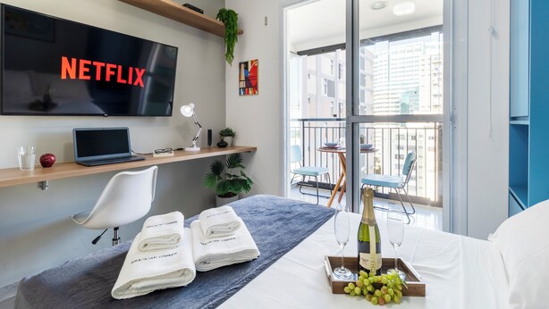A modern hotel room with a large TV displaying 'NETFLIX', a laptop, a desk with a plant, a chair, a bed with a white bedspread, a bottle of champagne, and a tray with grapes. - Vista Incrível com piscina e academia (Sao Paulo)