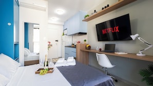 A modern hotel room with a bed, a desk with a laptop, a TV displaying 'NETFLIX', and a kitchenette in the background. - Vista Incrível com piscina e academia (Sao Paulo)