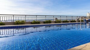 A rooftop pool with clear blue water, a metal railing, and a view of distant buildings. - Vista Incrível com piscina e academia (Sao Paulo)