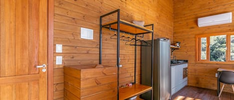 A wooden cabin interior with a kitchenette, a hanging clothes rack, and a window with a view of trees.