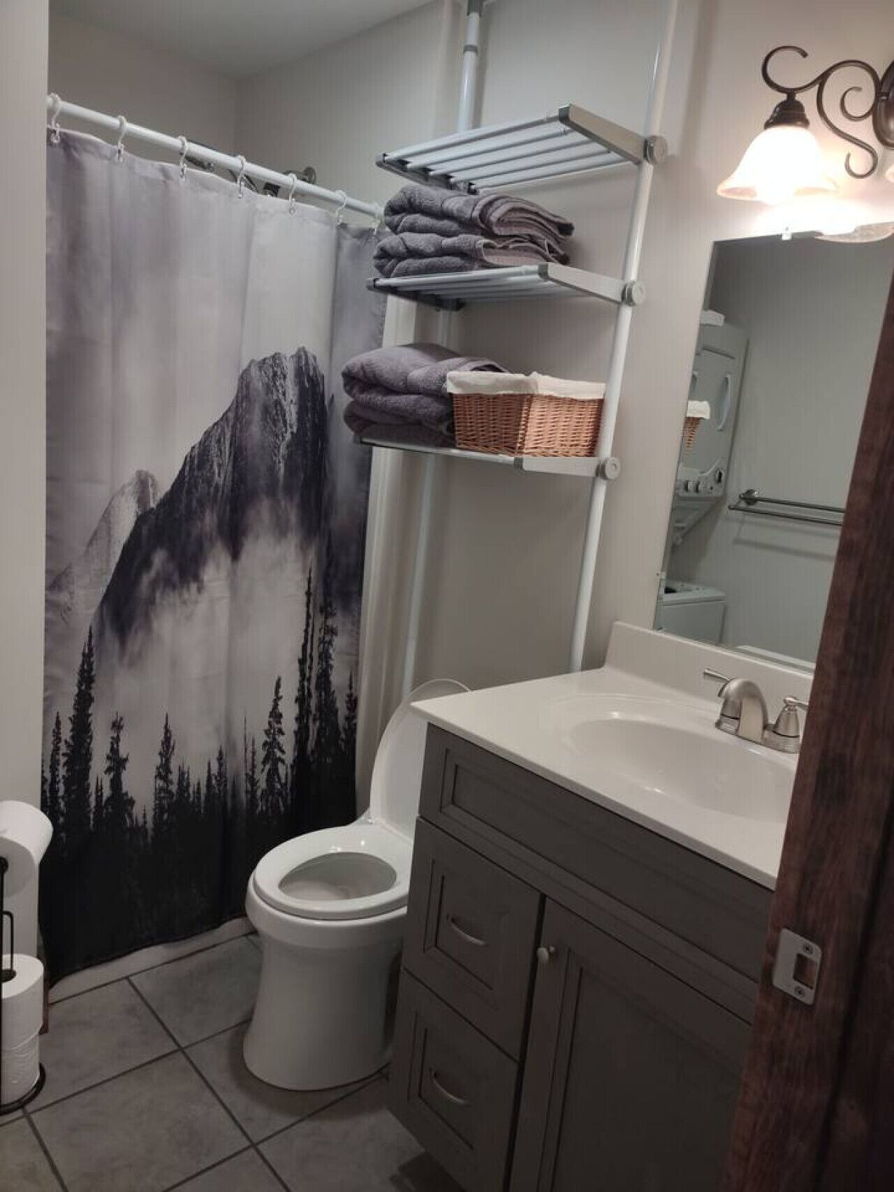 A bathroom with a mountain-themed shower curtain, a toilet, a sink with a mirror, and shelves with towels and a basket.
