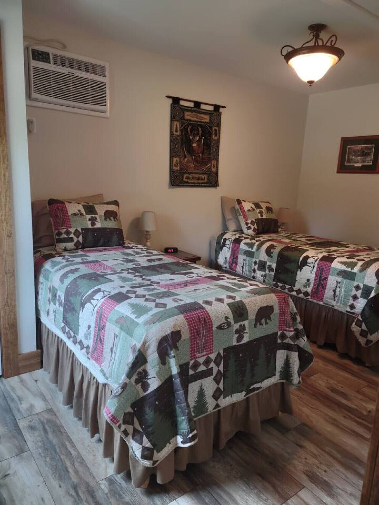 A room with two beds, each covered with a patterned quilt, a wall-mounted air vent, a decorative tapestry, and a ceiling light fixture.