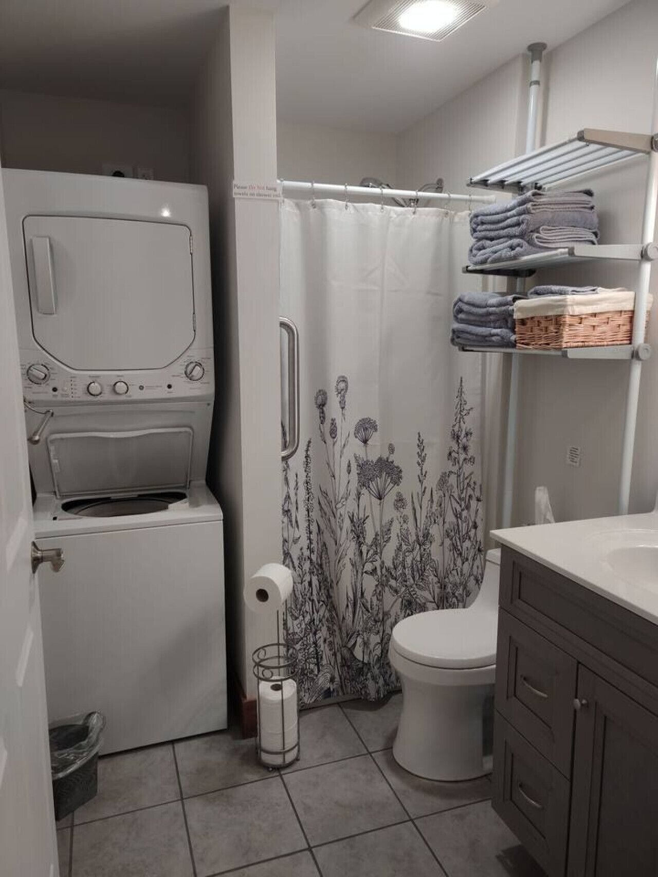 A compact bathroom with a washing machine, a toilet, a shower curtain with floral patterns, and a towel rack with folded towels.