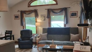 Una sala de estar acogedora con un sofá, un sillón y una mesa de centro. Hay un ventilador de techo, un televisor y una ventana con cortinas.
