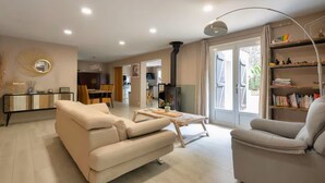 A modern living room with a sofa, armchair, and a coffee table. There is a bookshelf and a dining area visible in the background. - Villa 11 pers with jacuzzi - Lacanau, nature & ocean (Lacanau)