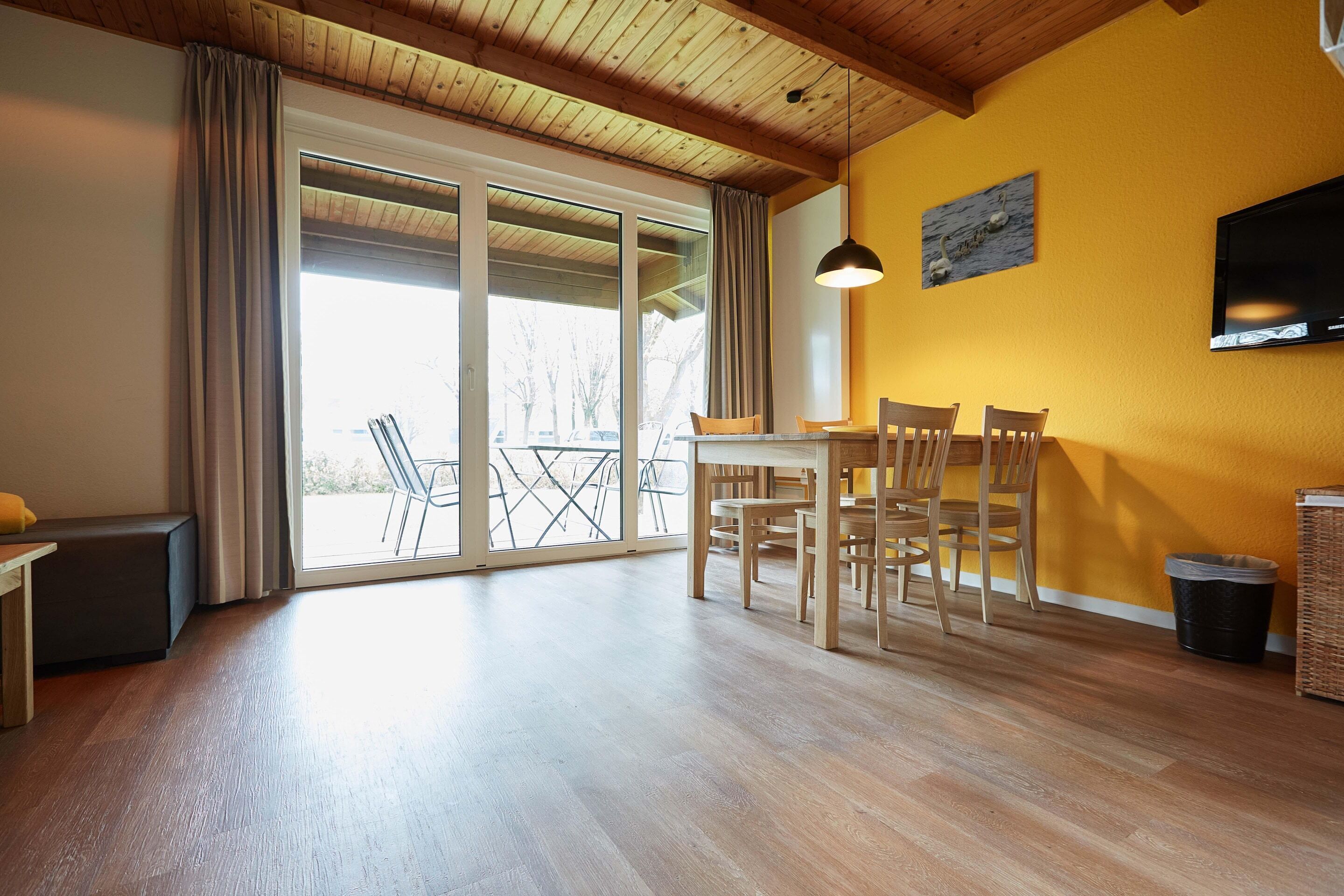 Een ruime kamer met houten vloer, een eettafel met stoelen, een bank, een televisie aan de muur en een grote glazen schuifdeur naar een buitenruimte.