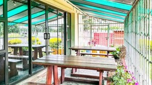 Ein überdachter Außenbereich mit Terrassentischen und Bänken, umgeben von Glaswänden und einem Metallzaun.