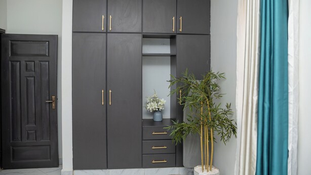A dark-colored wardrobe with gold handles, a small shelf with a vase of flowers, and a bamboo plant beside it. - Lovely apartment with WiFi in vibrant Kampala with a cozy and calm environment. (Kampala)