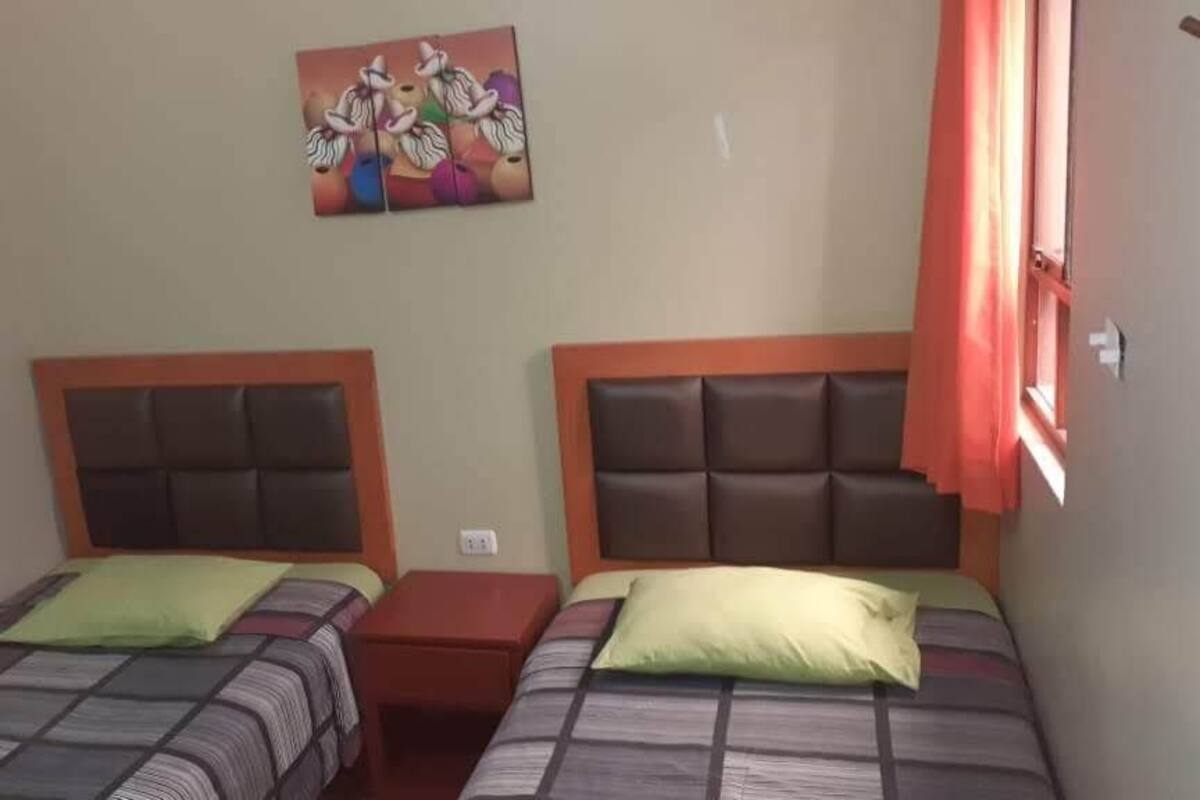 A room with two beds, each with a headboard and pillow. There is a small table between the beds, and a painting on the wall above the beds.